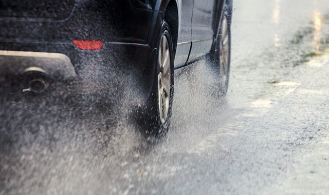 A dark SUV drives away from the camera through puddles of water, splashing the water into the air.